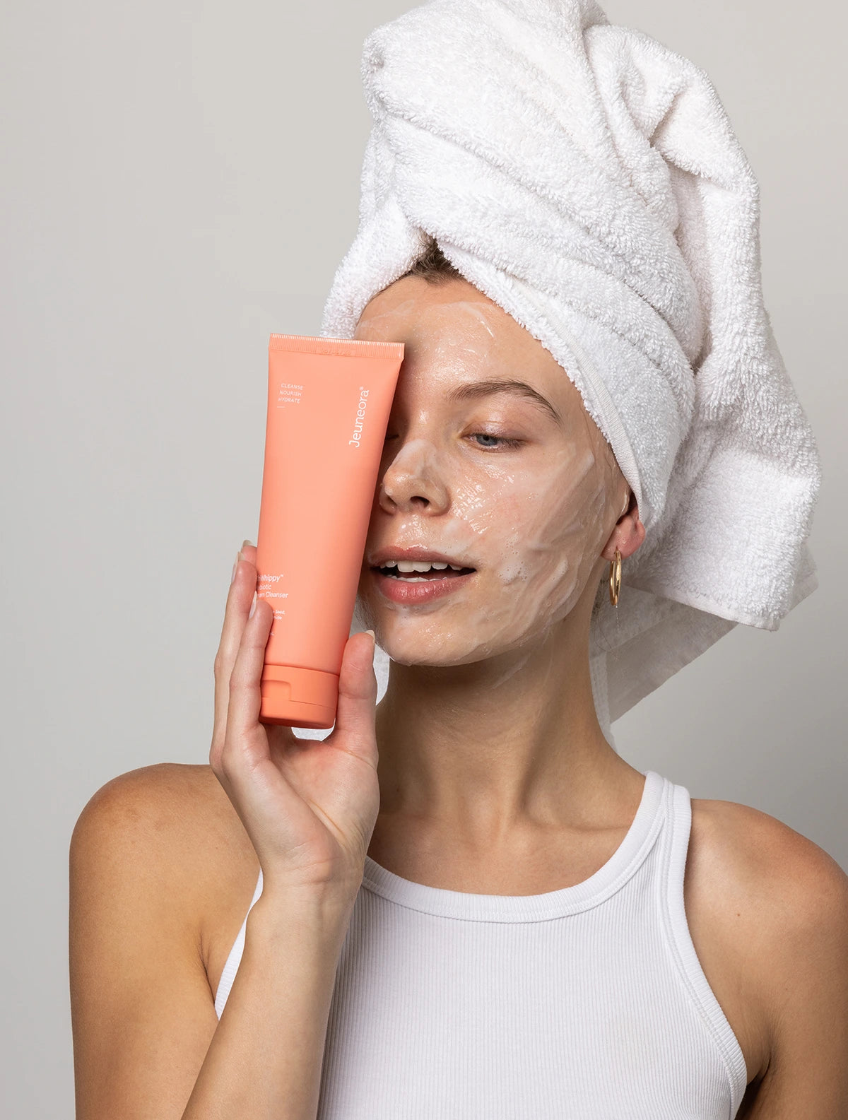 A woman with a towel on her head and cleanser on her face holds an OhWhippy Cream Cleanser