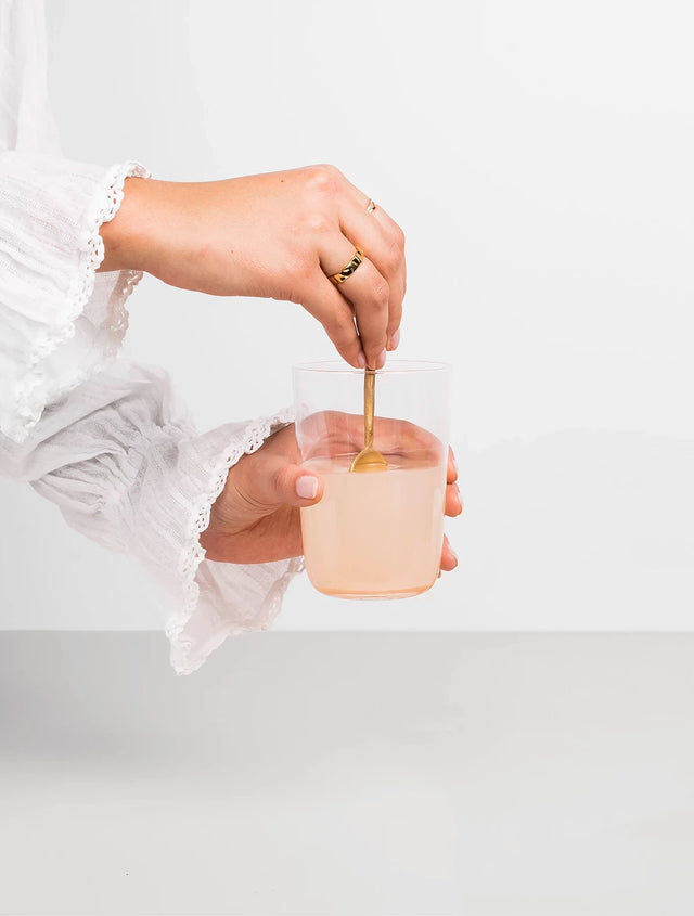 Woman stirring a glass of marine collagen drink with gold spoon.